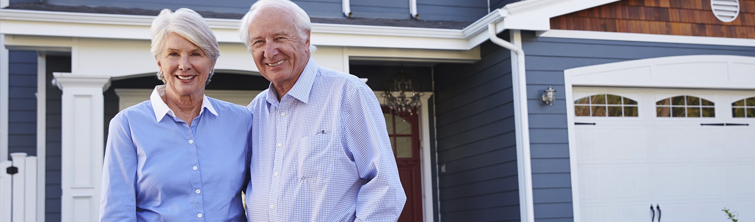 close-up of elderly couple smiling and standing in front of residence